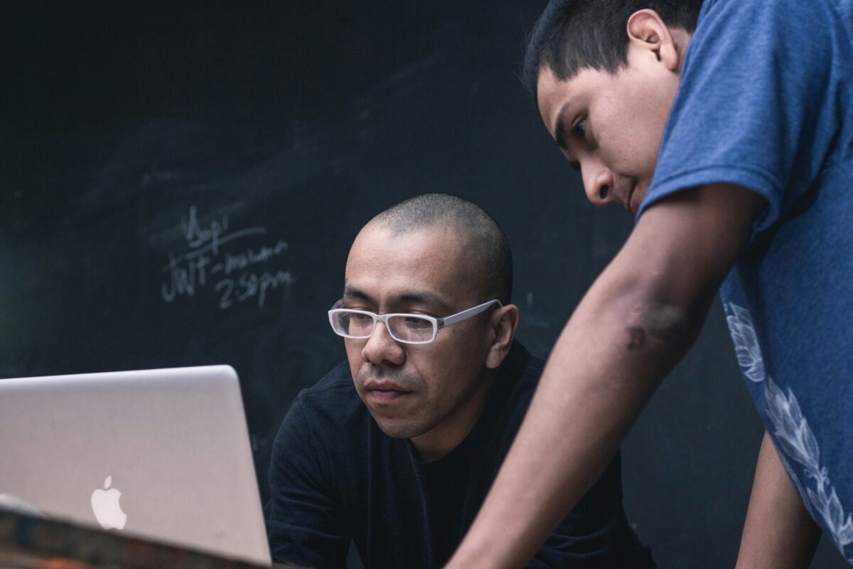 Dois estudantes se concentram em uma tela de computador, colaborando em um projeto em frente a um quadro negro com anotações. A imagem ilustra as dificuldades que muitos alunos cotistas enfrentam ao ingressar em universidades onde a maioria dos docentes é branca e as práticas institucionais muitas vezes não consideram suas experiências de vida. Esse ambiente pode gerar sofrimento psíquico e, em casos extremos, levar à evasão acadêmica.