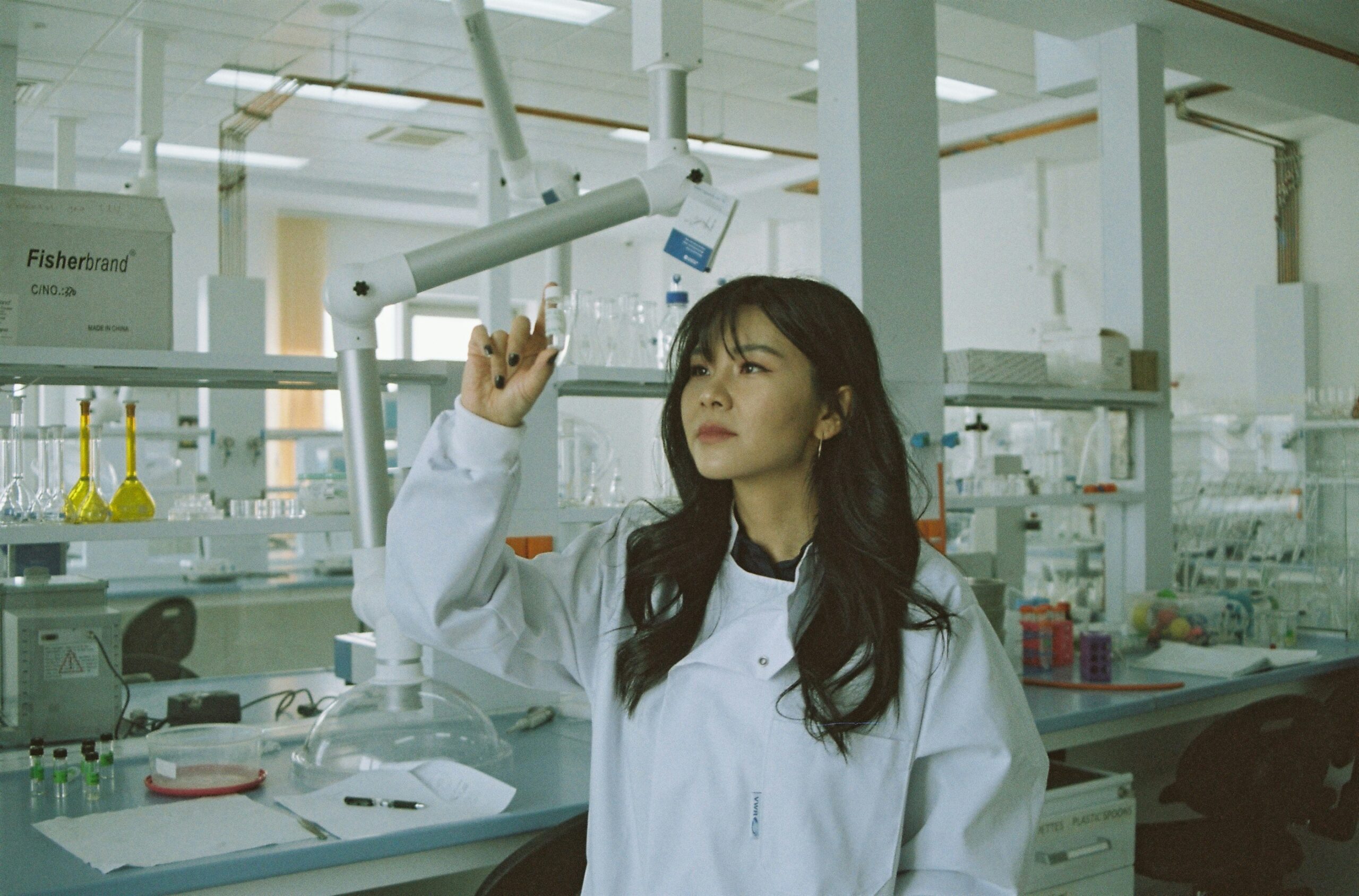 Jovem pesquisadora em um laboratório, vestindo jaleco branco, observa atentamente uma amostra que segura entre os dedos. Ao fundo, equipamentos de laboratório e frascos com líquidos coloridos compõem o cenário, refletindo o ambiente típico de pesquisa científica.