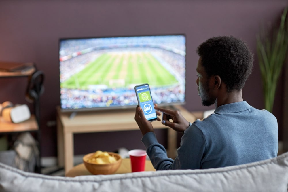 Rapaz sentado no sofá assistindo a um jogo de futebol na televisão e, usando um smartphone, acessa uma plataforma de apostas esportivas