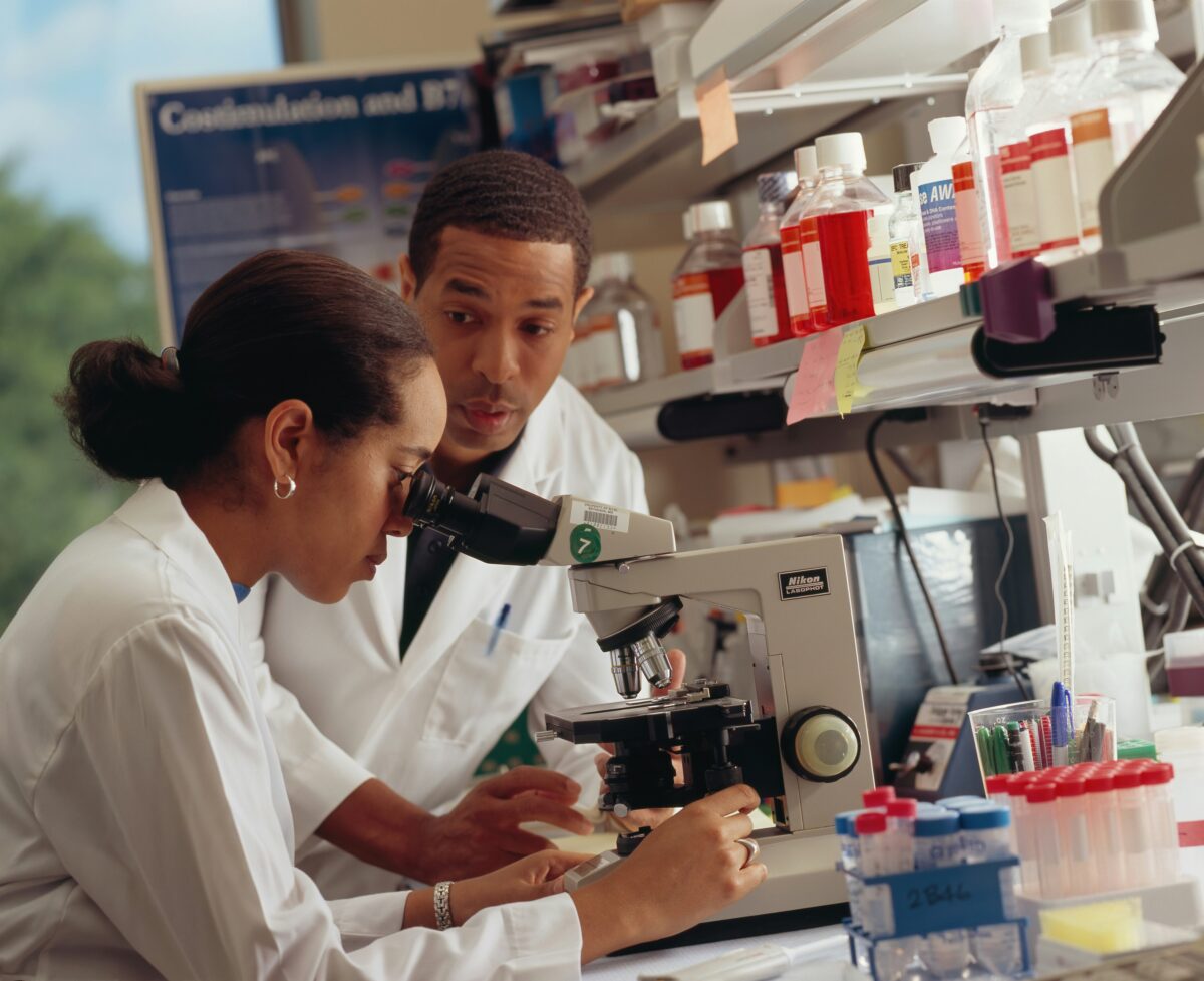 O Dois cientistas em um laboratório, um observando amostras no microscópio e outro acompanhando a análise, com frascos e materiais de pesquisa ao fundo.