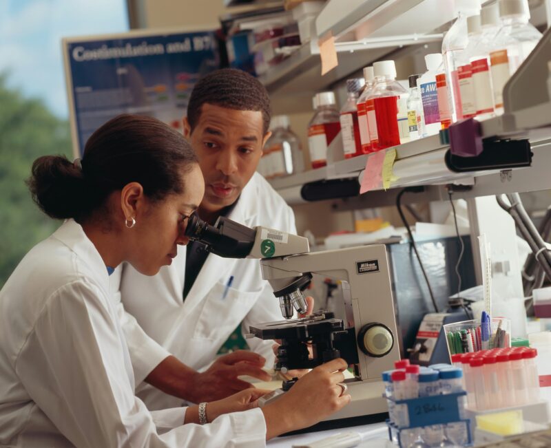 O Dois cientistas em um laboratório, um observando amostras no microscópio e outro acompanhando a análise, com frascos e materiais de pesquisa ao fundo.