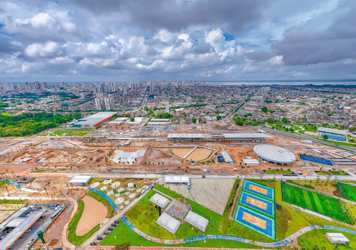 Vista aérea do Parque da Cidade, em Belém (PA), com obras em andamento. No primeiro plano, áreas de lazer com quadras esportivas, gramados e construções. Ao fundo, a zona urbana da cidade e o horizonte marcado por prédios e o rio. Céu parcialmente nublado.