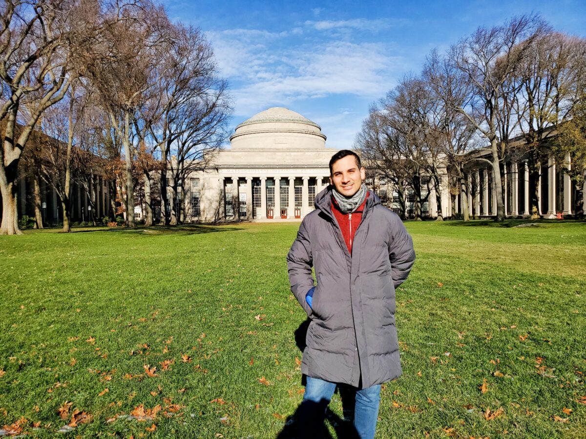 Daniel Dahis, in a gray winter coat, poses smiling on the grass before a neoclassical building, under a blue sky and with a backdrop of leafless trees