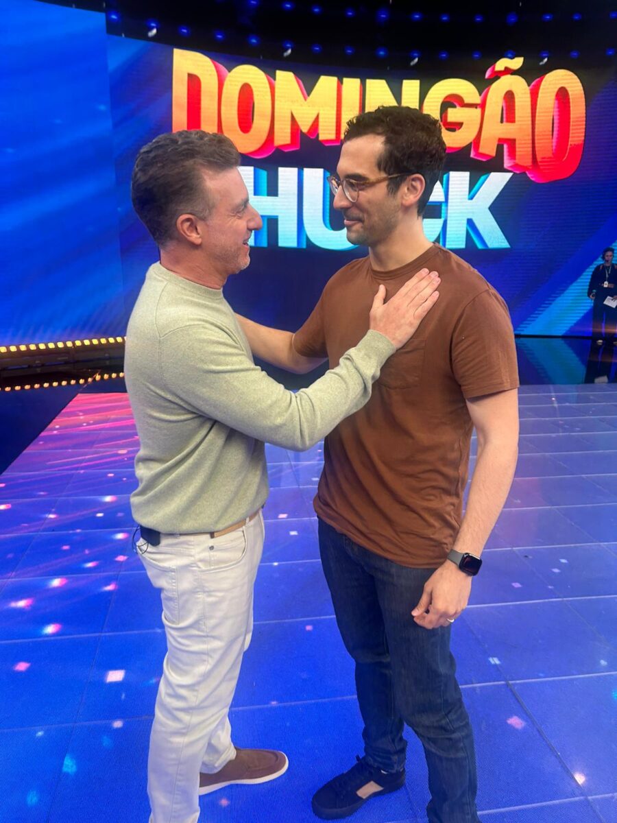 Daniel Dahis cumprimenta Luciano Huck no palco do programa Domingão com Huck; ambos sorriem e conversam lado a lado diante de um letreiro luminoso do programa.