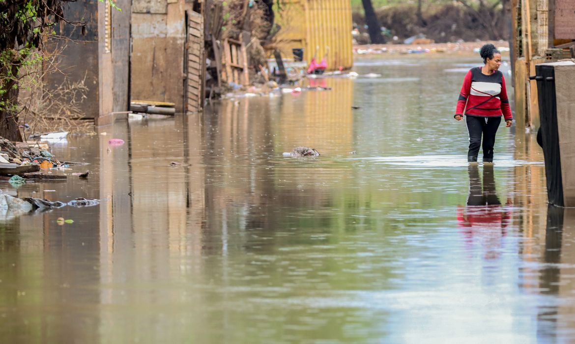 Mulher andando em rua inundada entre casas de madeira, com água até a canela, após enchente em comunidade vulnerável