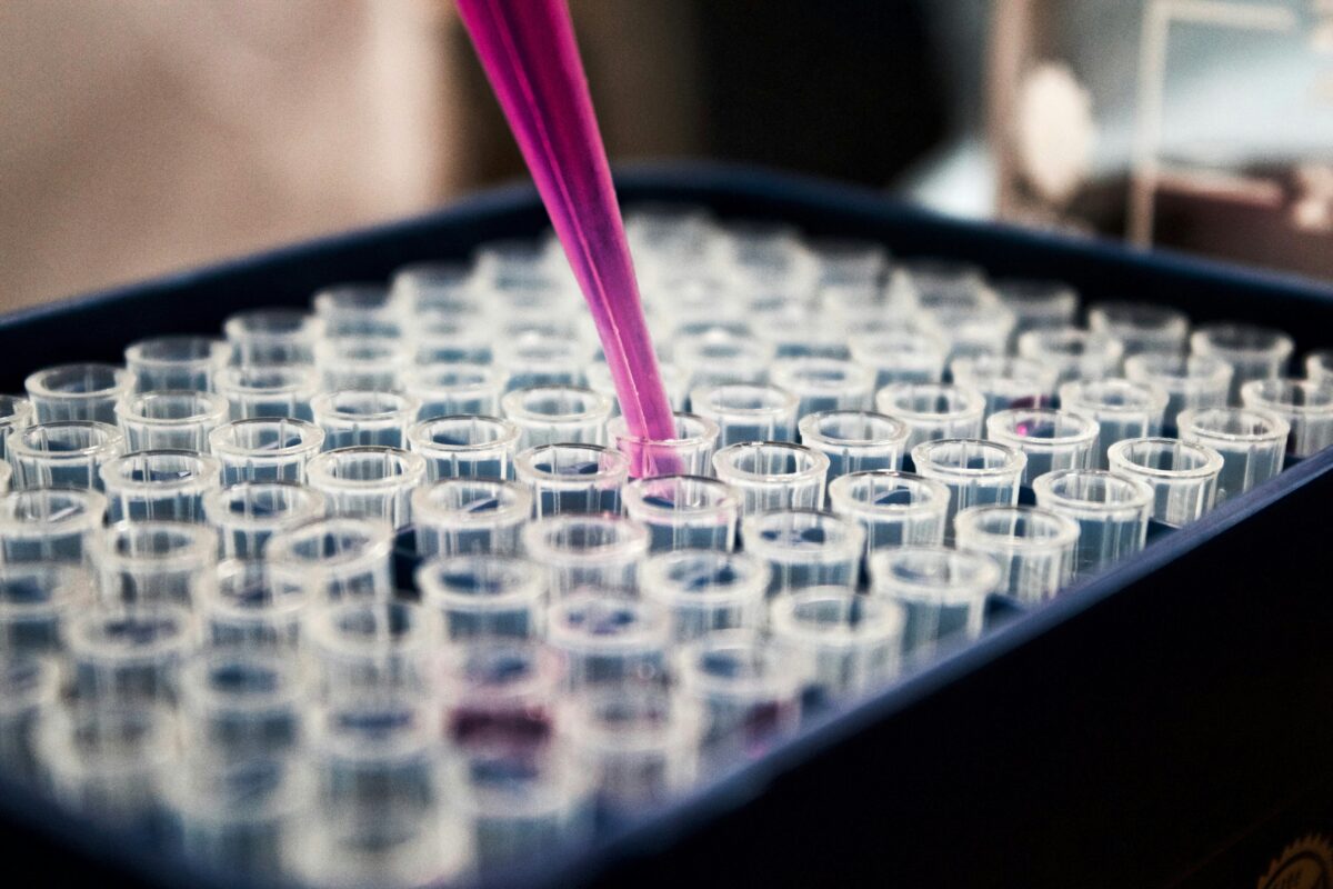 Pipeta deposita líquido rosa em uma das dezenas de microtubos organizados em uma bandeja de laboratório, representando o processo de análise em pesquisas clínicas