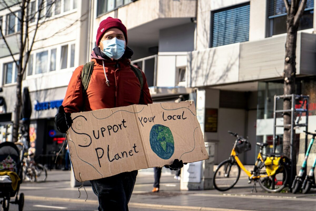 Pessoa usando máscara facial e touca vermelha segura um cartaz de papelão escrito "Support your local planet", com um desenho do planeta Terra. A cena ocorre em uma rua movimentada, com bicicletas, prédios residenciais e comércio ao fundo.
