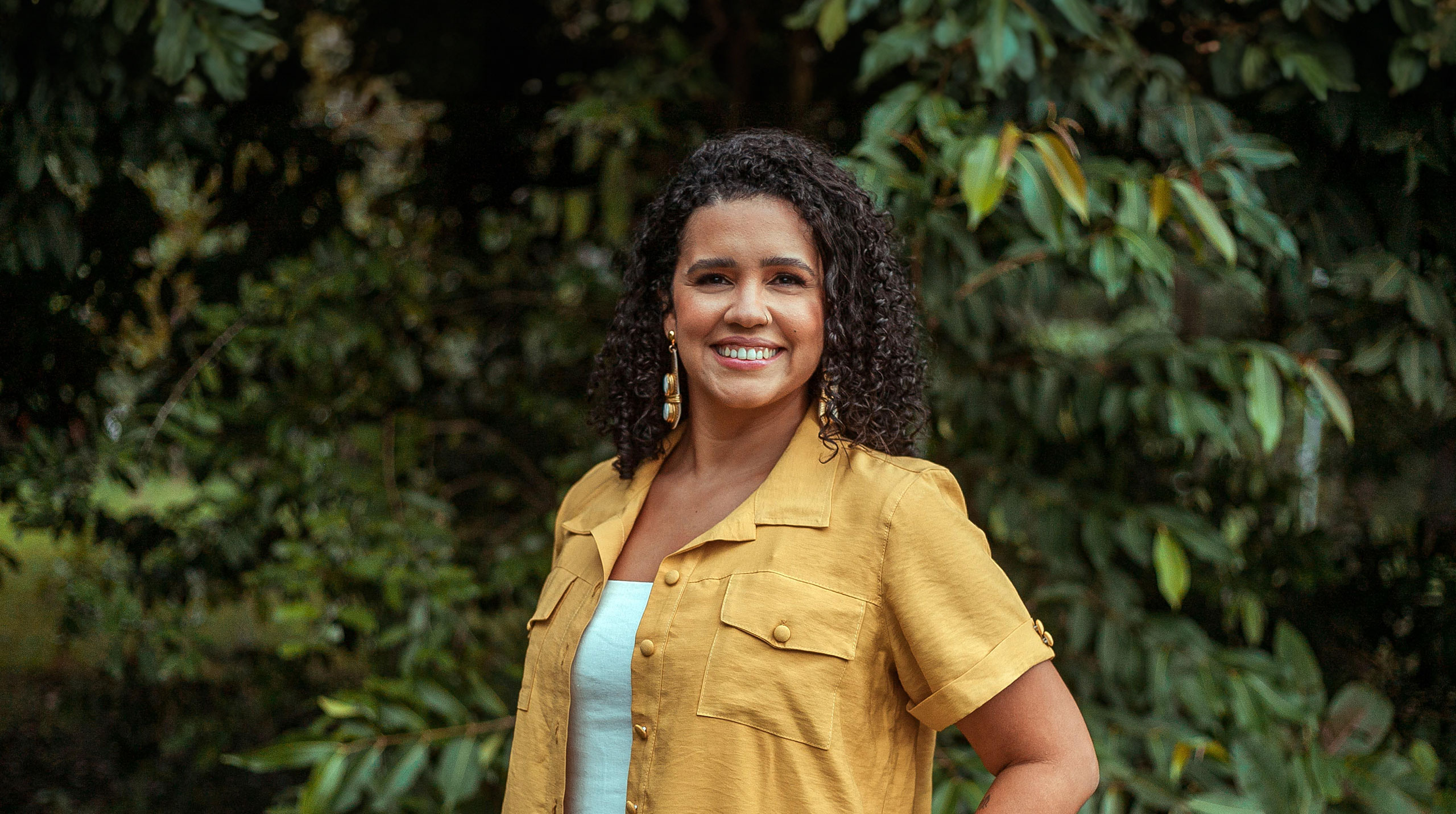 Mulher de cabelos cacheados e escuros sorri para a câmera em um ambiente externo com vegetação ao fundo. Ela veste uma camisa mostarda sobre uma roupa azul claro e posa com uma das mãos no bolso, transmitindo simpatia e confiança.