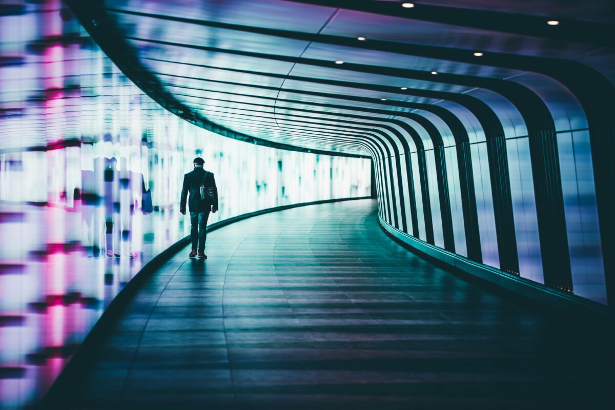 Pessoa caminha por um túnel em curva, futurista, iluminado por luzes coloridas, que leva a um lugar ainda desconhecido