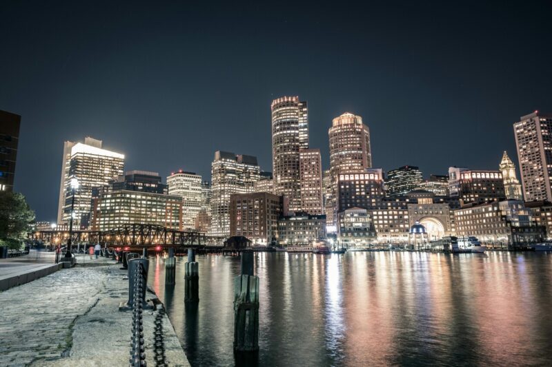 Vista noturna da orla de Boston, Massachusetts, com prédios iluminados refletidos nas águas do porto. A imagem mostra o skyline da cidade, que abriga importantes centros de pesquisa científica e instituições de ensino.