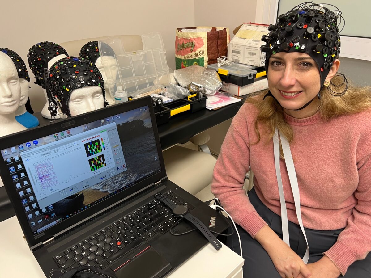 Eleonora Zioni sits in a lab next to a laptop, wearing a brain mapping helmet with sensors attached to her head, with equipment in the background