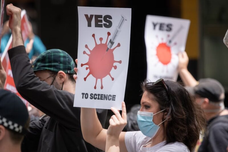 Mulher com máscara segurando cartaz com a frase "Yes to science" e a imagem de uma seringa apontada para um vírus estilizado, em manifestação a favor da ciência; outras pessoas com cartazes semelhantes aparecem ao fundo.