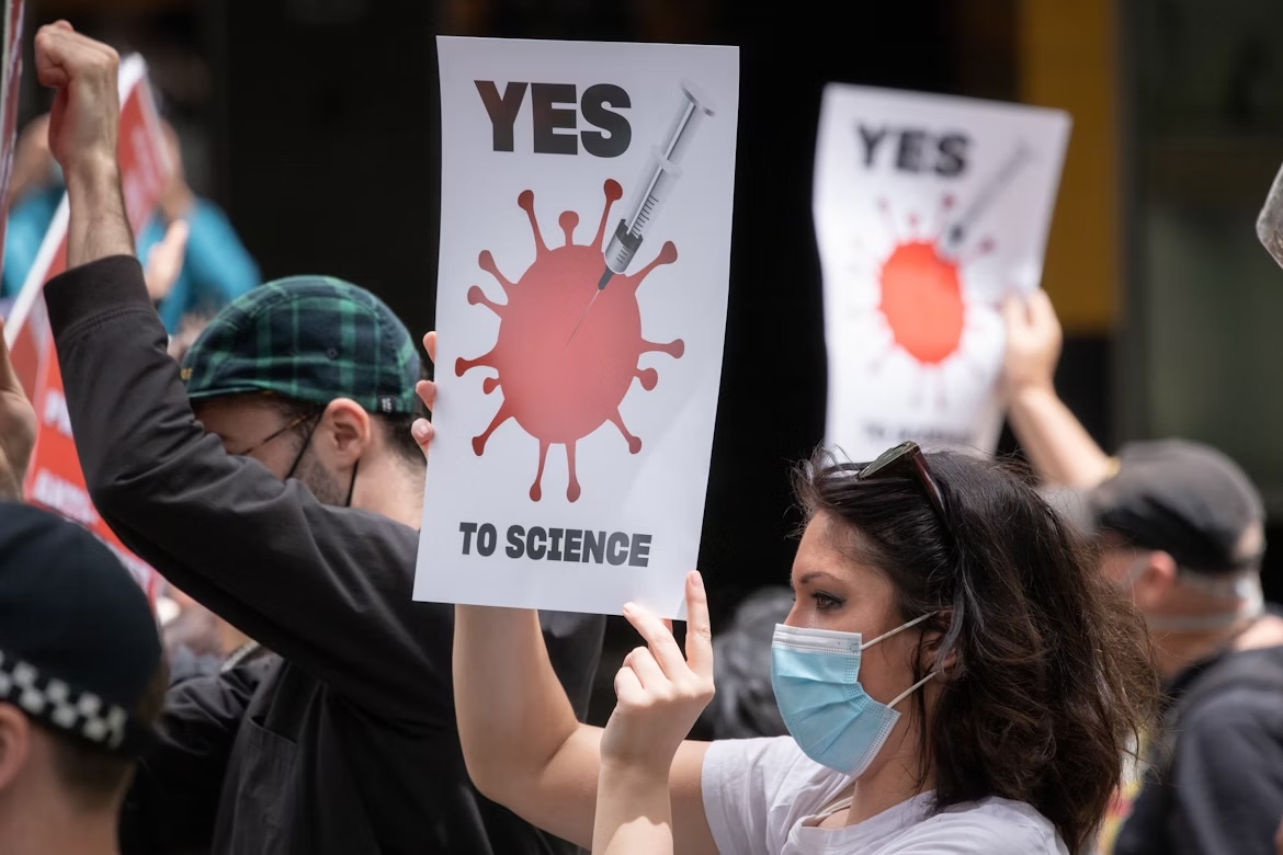 Mulher com máscara segurando cartaz com a frase "Yes to science" e a imagem de uma seringa apontada para um vírus estilizado, em manifestação a favor da ciência; outras pessoas com cartazes semelhantes aparecem ao fundo.