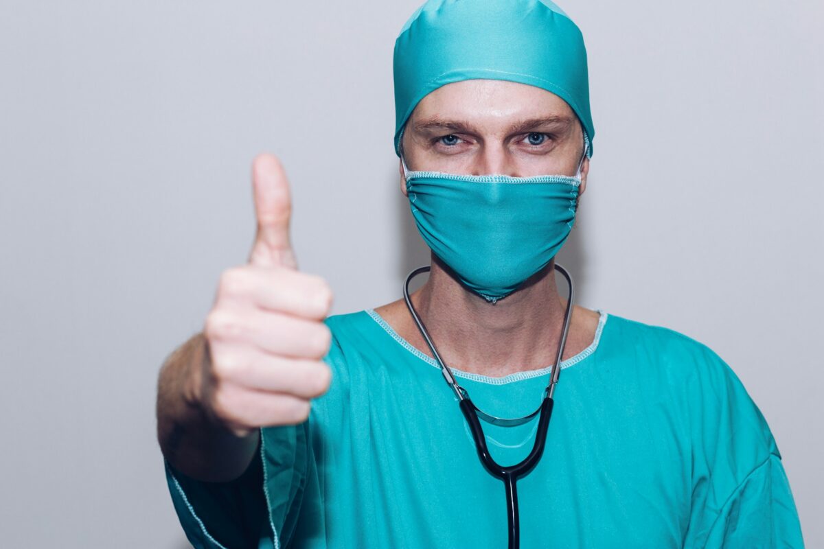 Médico com máscara cirúrgica, touca e estetoscópio fazendo sinal de "joinha" com a mão, em gesto de aprovação