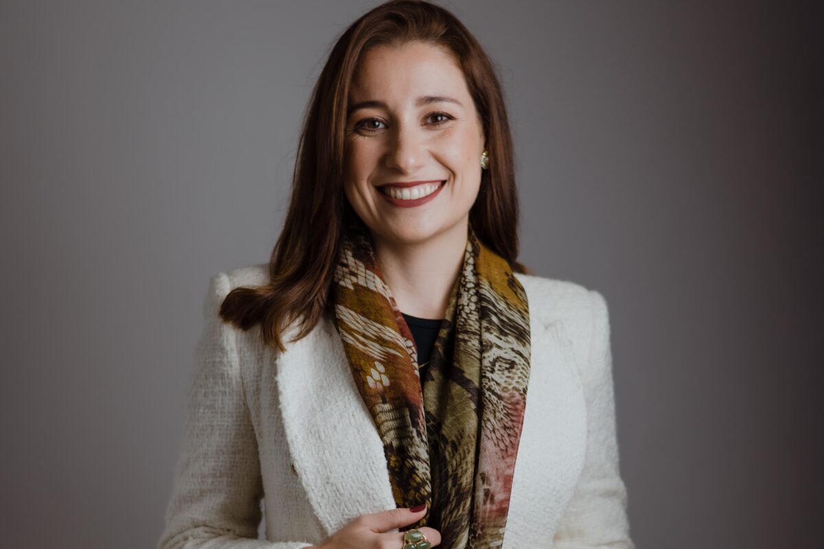 Jaqueline Ribas, an expert in recruiting research professionals, smiles at the camera, wearing a white coat and an earth-toned scarf, in front of a neutral background.