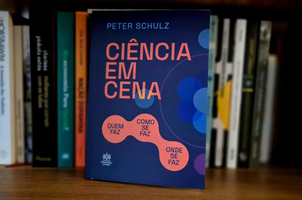 Fotografia do livro “Ciência em Cena”, de Peter Schulz, apoiado em uma estante com outros títulos ao fundo. A capa é azul com elementos gráficos circulares em tons de roxo e laranja, destacando as palavras Quem faz, Como se faz, Onde se faz.