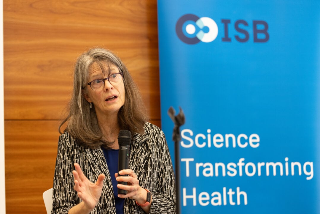 Mulher de cabelos grisalhos e longos, usa óculos e fala segurando um microfone durante uma coletiva de imprensa. Atrás dela há um painel azul com o logotipo do Instituto de Biologia de Sistemas (ISB) e o texto “Science Transforming Health”.