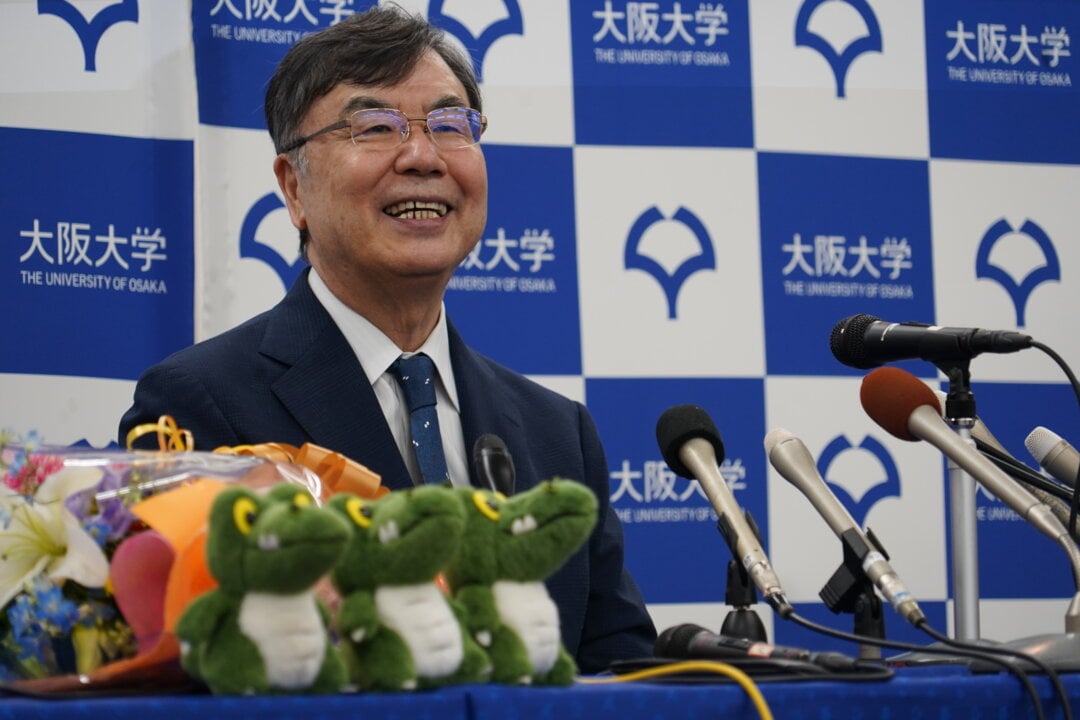 Homem asiático de terno azul e gravata, sorri durante uma coletiva de imprensa na Universidade de Osaka. À frente dele há microfones, flores e pelúcias de sapos; ao fundo, um painel com o logotipo e o nome da universidade em japonês e inglês.