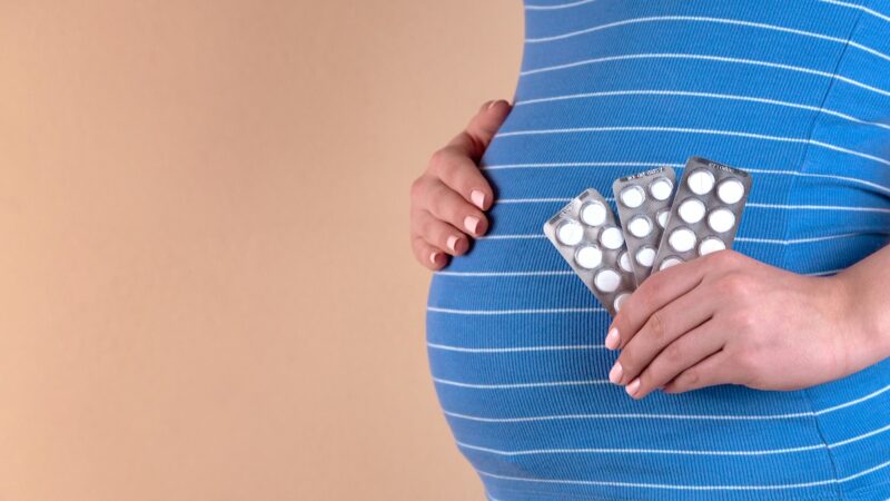 Pregnant woman with a blue striped blouse holds three packs of white pills on her abdomen, illustrating the use of medications during pregnancy