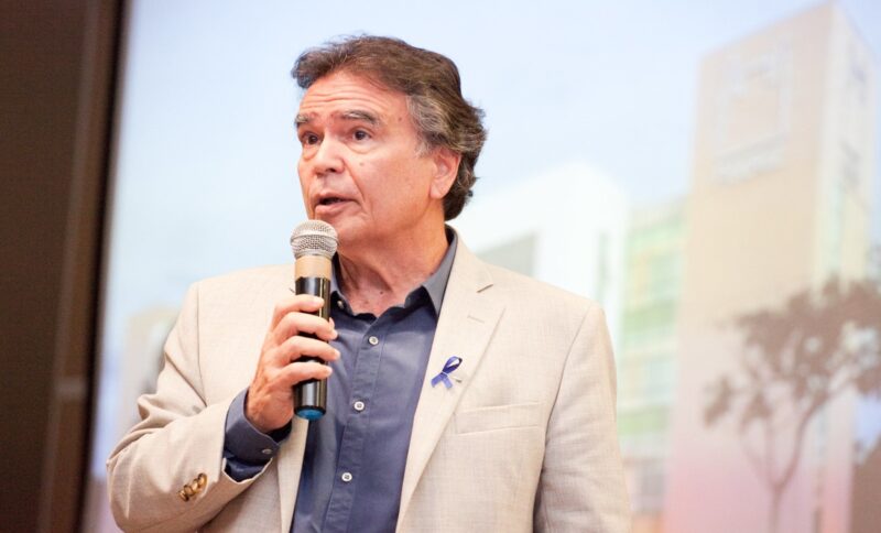 The image shows public health physician José Gomes Temporão, a middle-aged man wearing a light-colored blazer and a blue shirt, speaking into a microphone in an auditorium. A small blue ribbon is pinned to his jacket pocket. In the background, the light-colored interior of the building appears blurred.