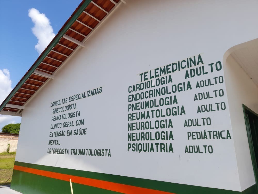 The image shows the white façade of a health facility, with the available medical services painted in green letters on the wall. On the left are specialties such as gynecology, rheumatology, general practice, mental health, and orthopedics. On the right is a list of telemedicine services, including cardiology, endocrinology, pulmonology, rheumatology, adult and pediatric neurology, and psychiatry. A wooden roof and blue sky complete the exterior scene.