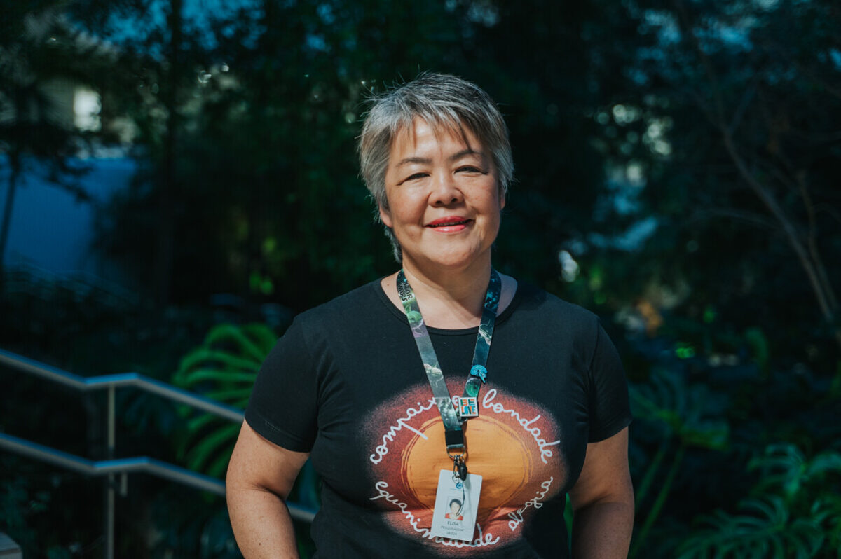 Elisa Kozasa é uma mulher de meia-idade, de cabelo curto grisalho, que na imagem sorri levemente para a câmera, usando camiseta preta com estampa colorida circular e crachá pendurado no pescoço, em ambiente externo com vegetação ao fundo 