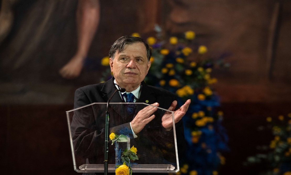 Physicist Giorgio Parisi appears wearing a dark suit, speaking from behind a transparent lectern decorated with yellow roses. Behind him, a dark panel and a blurred arrangement of yellow and blue flowers.