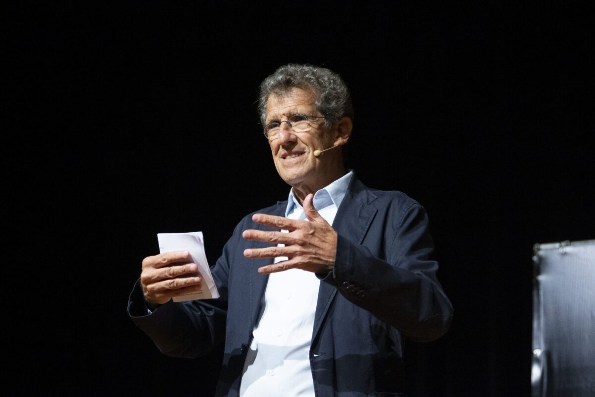 USP Professor Carlos Monteiro on a darkened stage, wearing a suit jacket and light-colored dress shirt, speaking to the audience and gesturing with his hands while holding notes in one hand.