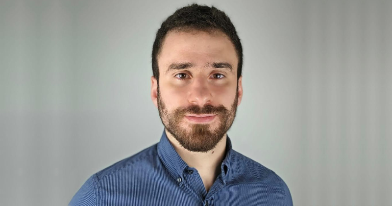 Retrato de Leonardo Saraiva, um jovem homem com barba curta, cabelos curtos e camisa social azul, posando para a câmera com fundo neutro