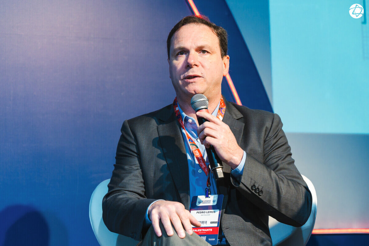 Lemos, wearing a suit and seated in an armchair on a stage, speaks into a microphone at a conference. The atmosphere is professional, with stage lighting and a setting typical of a scientific event.