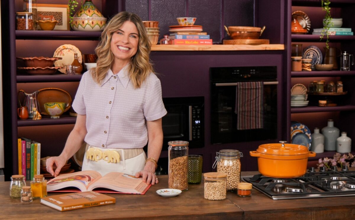 Rita Lobo in a colorful kitchen, leafing through an open cookbook on a wooden countertop. In the background, pots, dishes, and cookbooks are arranged on purple shelves. An orange cast-iron pot is on the stove, beside jars of grains and spices.