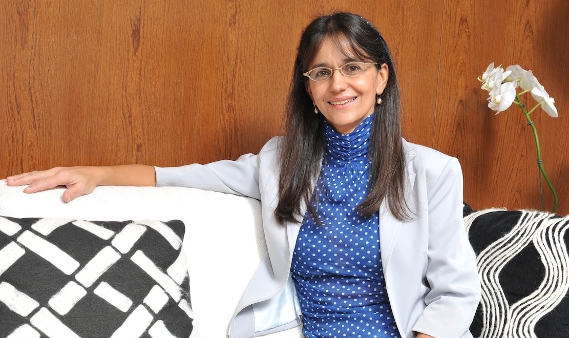 Photo of Sonia Mansoldo Dainesi, seated on a sofa in an indoor setting, with a relaxed posture and a calm expression. She wears light-colored clothing with blue tones, conveying professionalism and warmth. Discreet décor in the background creates a sober, contemporary atmosphere.