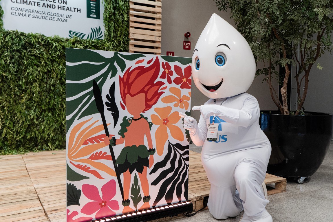 Mascot Zé Gotinha, wearing a white uniform and a SUS badge, poses alongside a colorful panel with an Indigenous illustration and floral elements during BHAP Day at the 2025 Global Conference on Climate and Health in Belém
