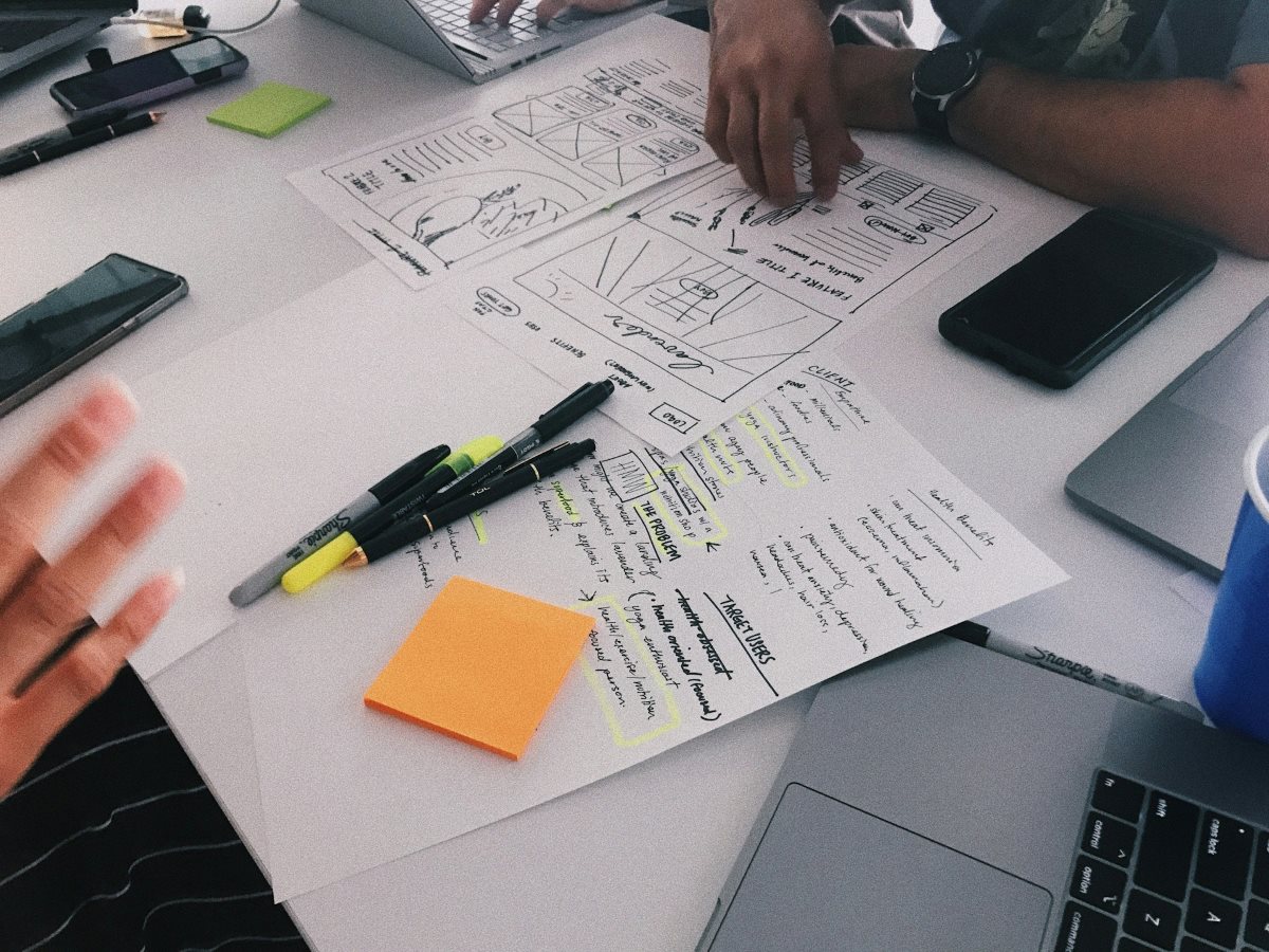 Worktable scattered with sketches, charts, and notes as team members discuss and point to documents. The scene conveys collaboration, strategic planning, and project development