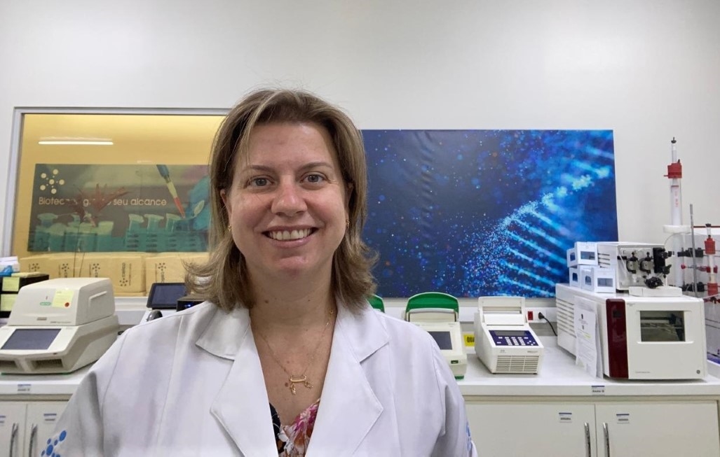 Pesquisadora sorridente veste jaleco em laboratório equipado com aparelhos científicos, tendo ao fundo uma imagem estilizada de DNA em tons de azul