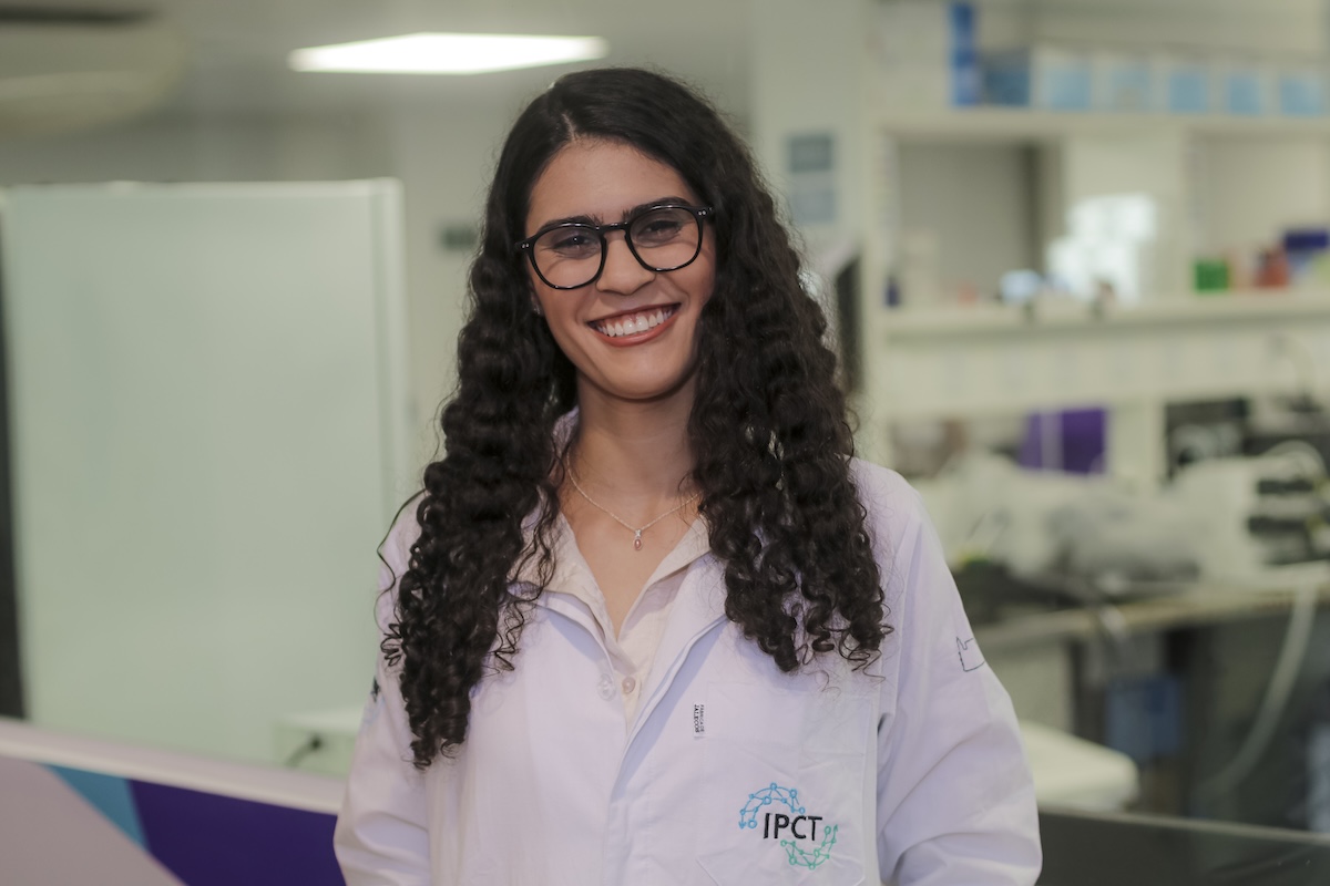 Beatriz Barreto-Duarte, mulher jovem de cabelos cacheados longos e óculos de armação preta, sorri para a câmera vestindo jaleco branco com logo do IPCT em laboratório com equipamentos ao fundo