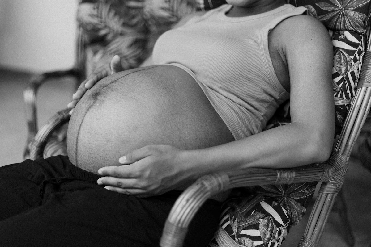 Black and white photograph of a pregnant woman sitting in a wicker chair with her hands resting affectionately on her belly. The close-up composition shows the pregnant woman's torso, highlighting her pregnant belly visible below a light-colored top. Blurred foliage in the background suggests a domestic setting with plants, conveying an atmosphere of intimacy and hope