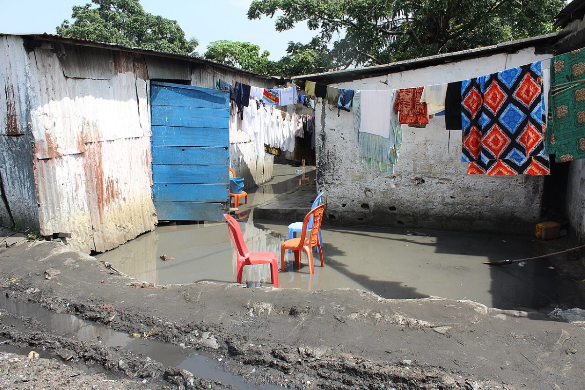 A flooded residential area composed of simple housing, clothes hanging out to dry, and partially submerged plastic chairs. The scene demonstrates the impacts of floods and poor sanitary conditions on the daily lives of the population.
