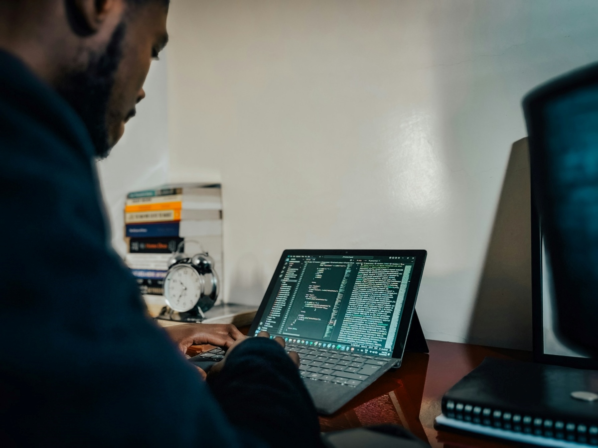 A man in an office works on a laptop with code and text on the screen, with books and a clock in the background. The image portrays the use of digital technologies and programming in the context of research and data analysis.