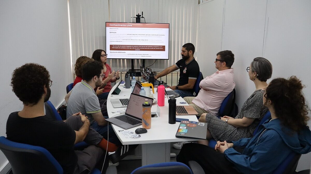 Two people sit at a table during a lecture in a classroom. One speaks into a microphone while the other listens attentively. On the table are glasses, a jug of water, and papers, and a chalkboard can be seen in the background.