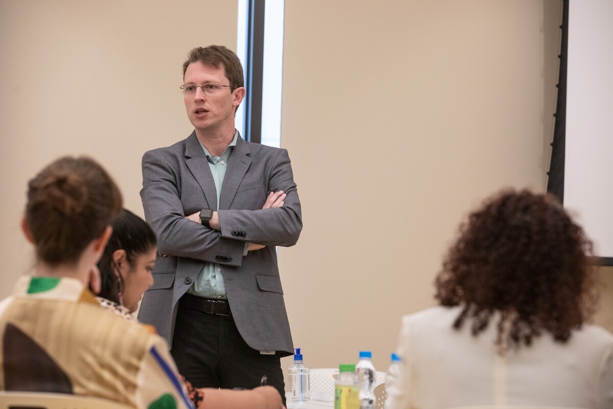 A man in a suit and glasses speaks while standing up, with his arms crossed, during a meeting or class, while three people sit at a table listening to him