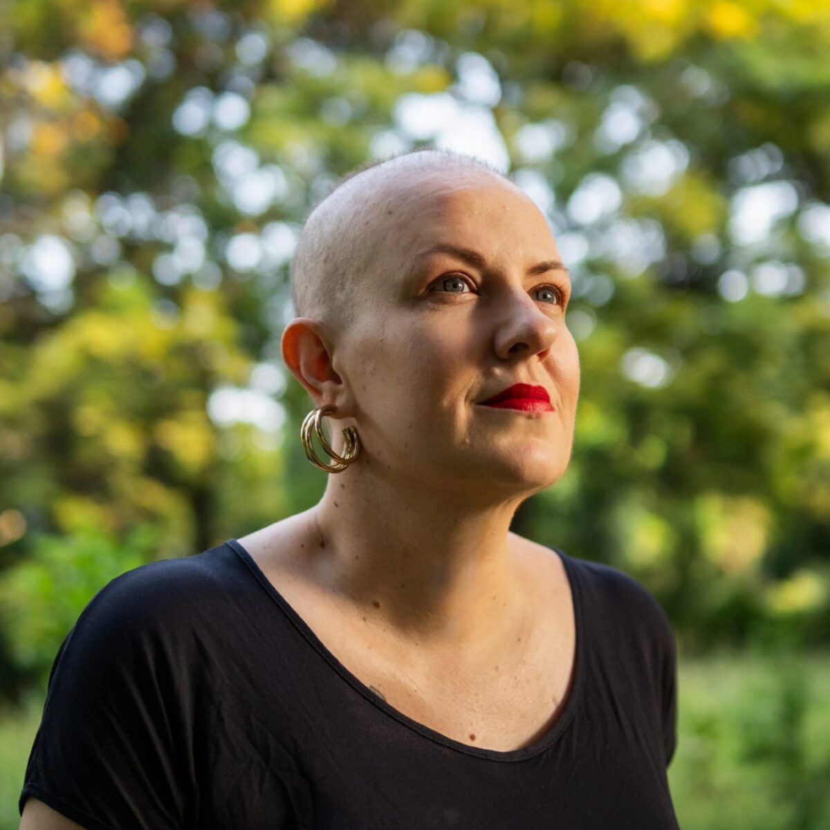 A woman with a shaved head, wearing gold earrings and red lipstick, looks at the horizon with a serene and determined expression. In the background, natural light filters through the green vegetation, creating a calm and contemplative atmosphere.