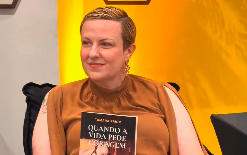 Writer Tamara Prior is seated in a studio with warm amber lighting in the background, holding the book Quando a Vida Pede Coragem. She is wearing a brown blouse and smiling slightly as she shows the book’s cover to the camera.