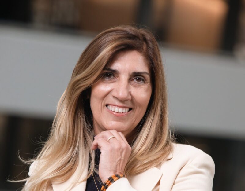 Texto Alternativo Retrato de mulher sorridente com cabelos castanhos longos, usando blazer branco sobre blusa preta