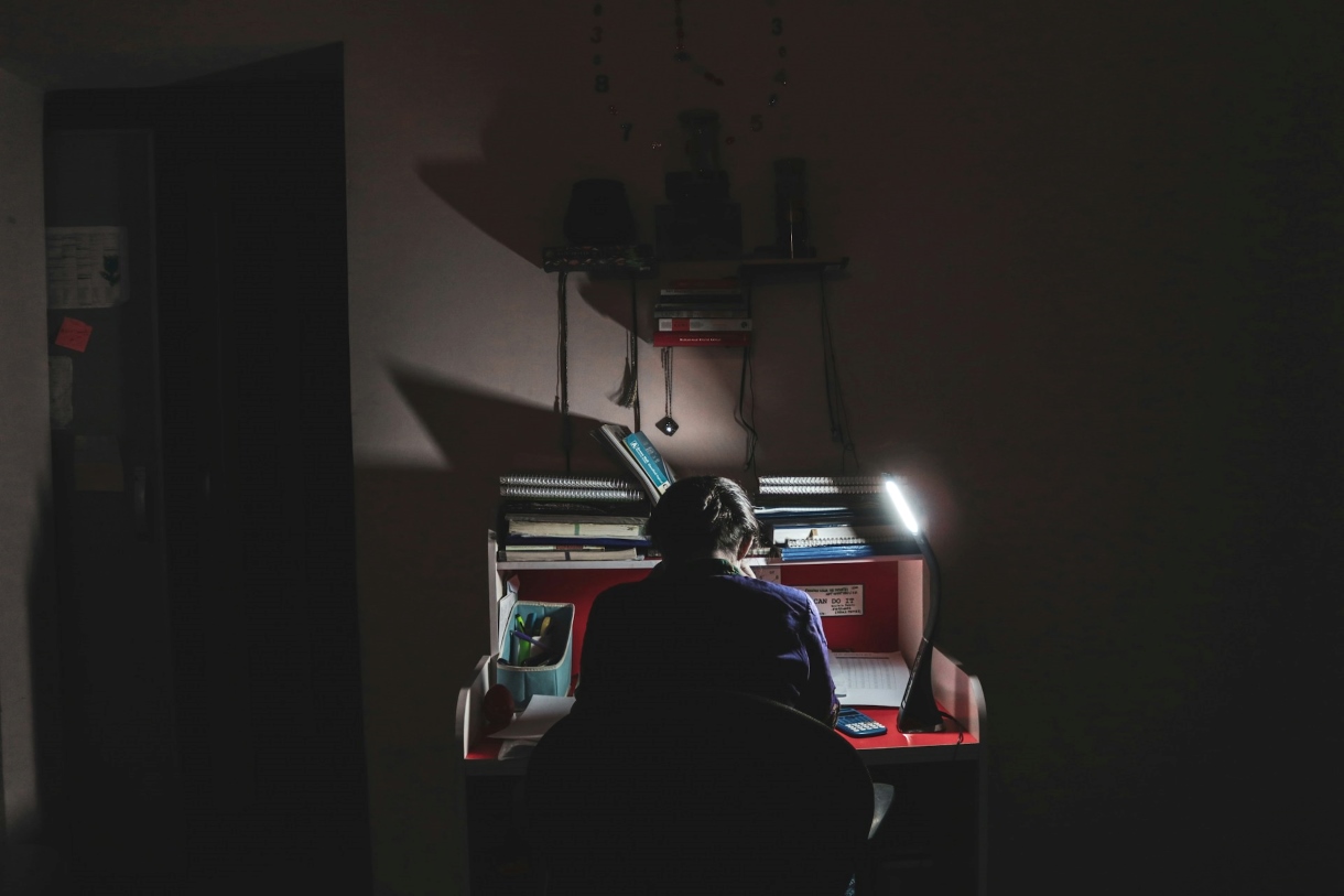 A person sitting at a desk in the dark, illuminated only by a lamp. The scene conveys intense concentration and a sense of isolation while working into the night.