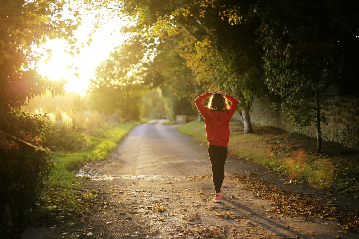 A person walking along a tree-lined road at dusk, illuminated by the golden light of the setting sun. The image conveys a sense of tranquility, movement, and connection with nature.