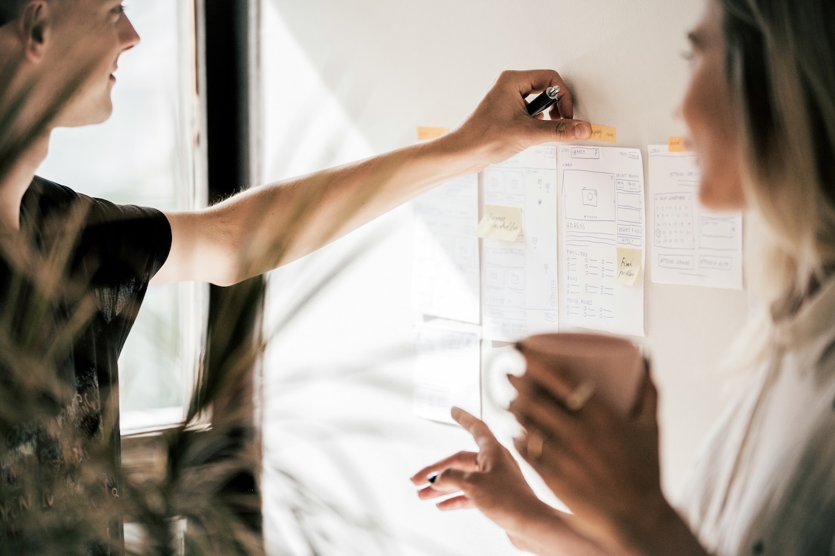 Two people collaborate in front of a board with wireframes and Post-it Notes, planning a project interface.