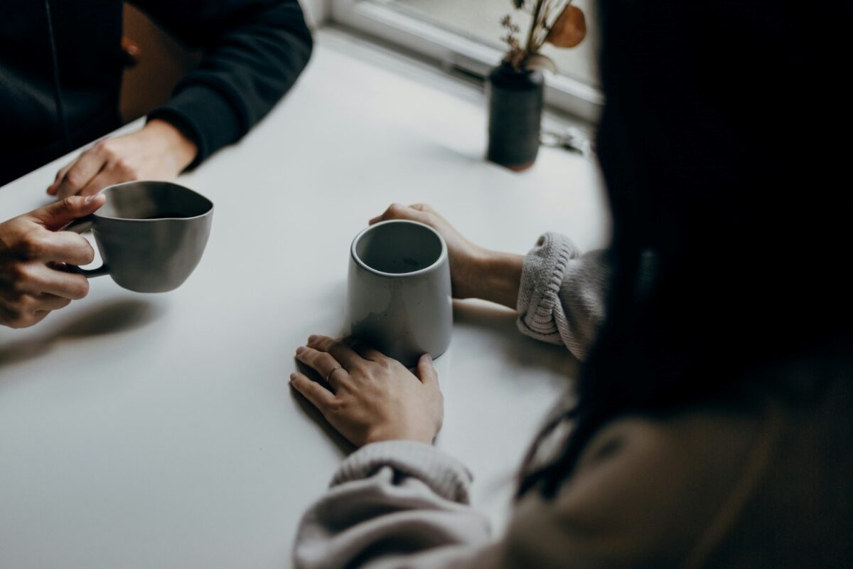 Two people sit at a table, holding mugs, engaged in intimate conversation. Soft lighting and close-up framing suggest a friendly atmosphere and attentive listening