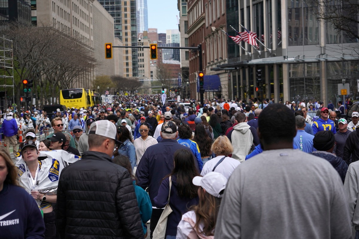 Large numbers of people walking on a wide urban avenue on a sunny day. In the background, commercial buildings, traffic lights, and sidewalks.