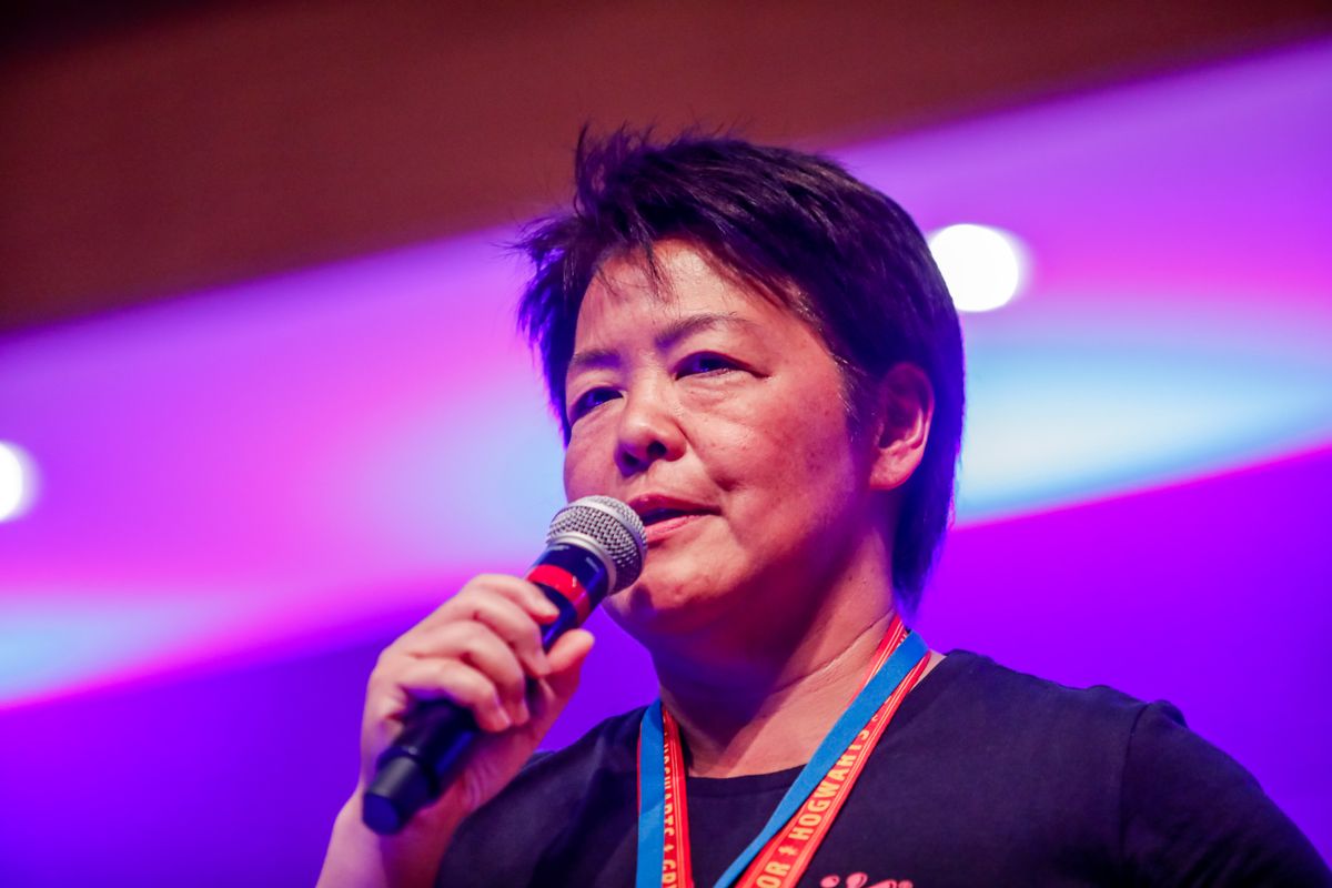 A pesquisadora Elisa Kozasa segura um microfone e fala ao público em um palco iluminado por luzes roxas e rosas.Ela usa crachá e camiseta escura, em contexto que sugere evento ou conferência científica